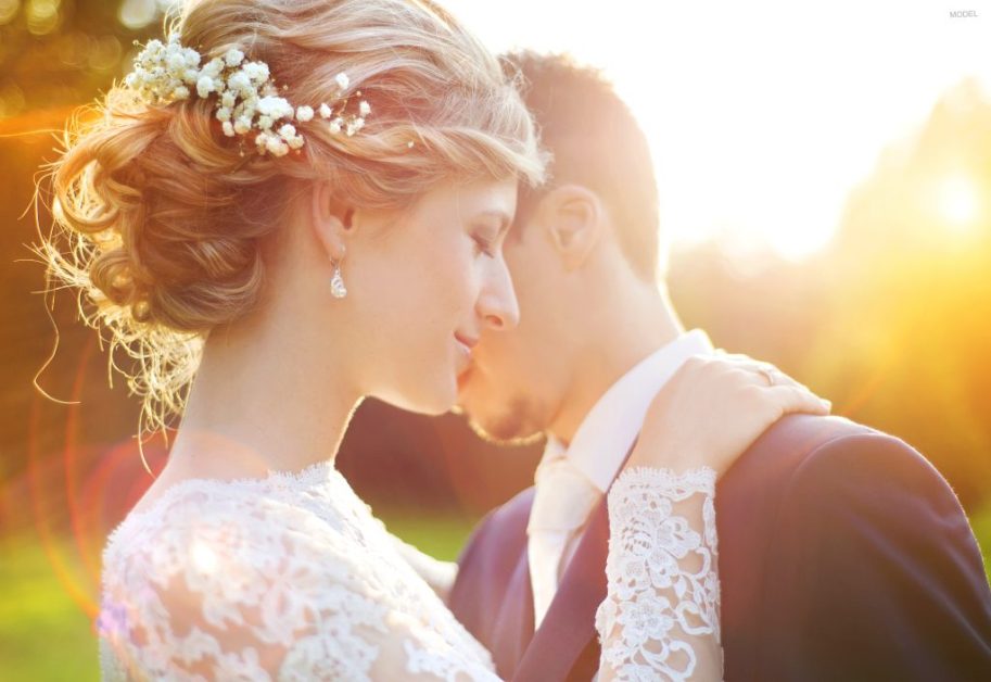 Bride and groom rest their faces against one another with the setting sun casting a gold glow on the trees behind them.