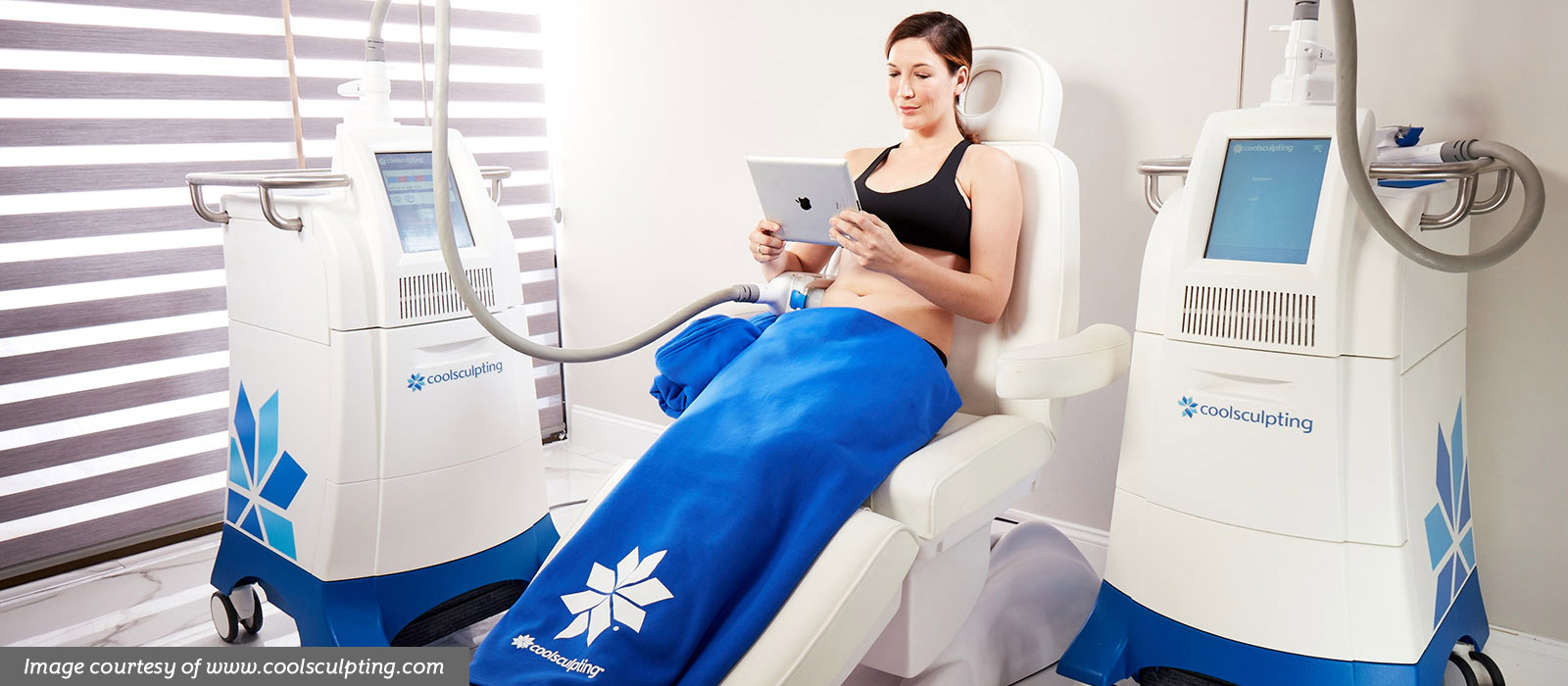 Woman in black sports bra reclines in treatment chair, reading an iPad, while a CoolSculpting paddle is attached to her abdomen