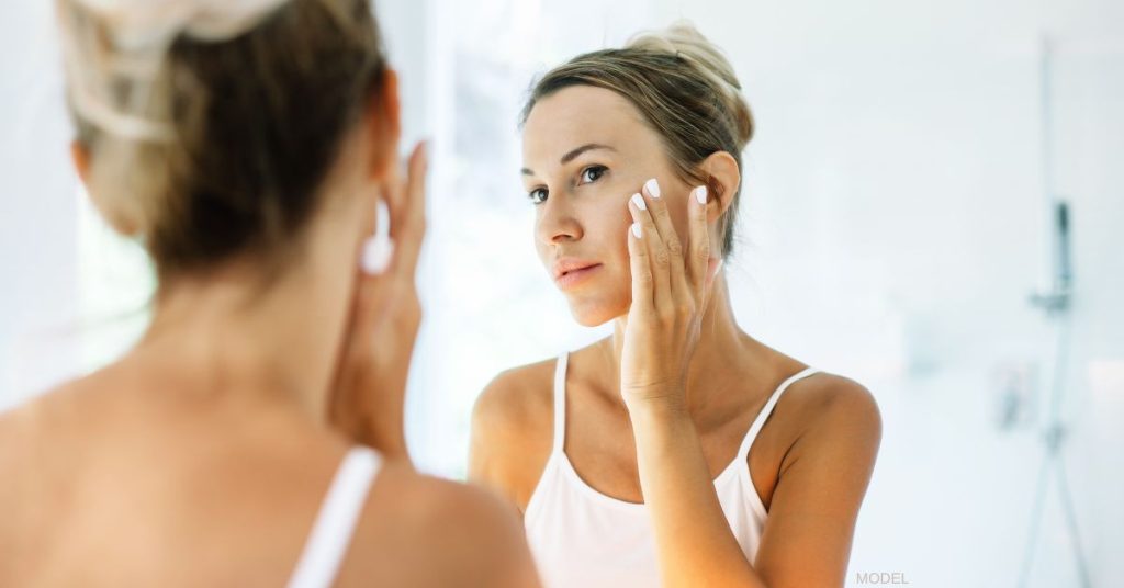 Woman looking at reflection in mirror and touching her cheek (model)